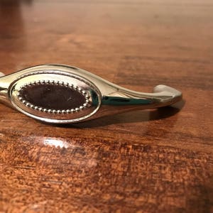 Może przedstawiać: Srebrny, owalny uchwyt do szafki z ozdobnym krawędzią i mniejszym, owalnym wkładką w środku.