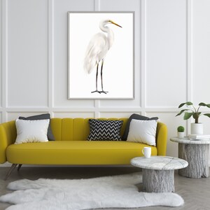 May include: A white egret bird stands on a white background in a framed print. The print is hanging on a white wall above a yellow sofa with pillows. There is a small plant and a side table with a book on it to the right of the sofa.