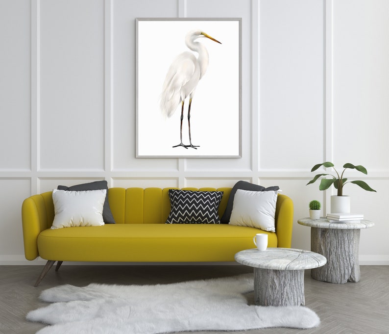 May include: A white egret bird stands on a white background in a framed print. The print is hanging on a white wall above a yellow sofa with pillows. There is a small plant and a side table with a book on it to the right of the sofa.