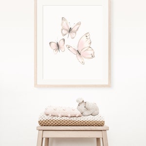 May include: A framed print with a white background featuring three pink butterflies in flight. The print is hanging on a white wall above a wooden stool with a knitted blanket and a knitted bunny toy.