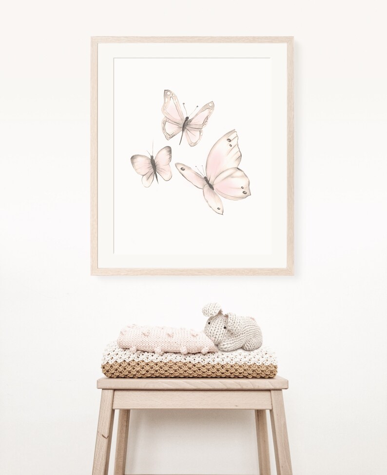 May include: A framed print with a white background featuring three pink butterflies in flight. The print is hanging on a white wall above a wooden stool with a knitted blanket and a knitted bunny toy.