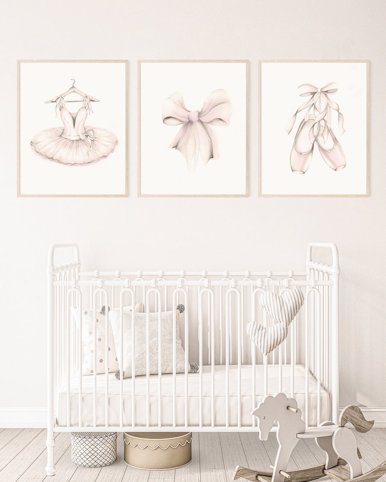 Set of three ballerina style unframed prints drawn in pencil with sepia and blush pink accents. The set includes a ballerina tutu, a pink bow and a pair of ballet slippers. The pictures are shown hanging on a baby girl's nursery wall.