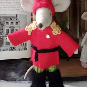 May include: A white mouse wearing a red and gold uniform with a black belt and black boots. The mouse is wearing a red hat with gold trim. The mouse is standing on a book.