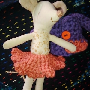 May include: A handmade stuffed mouse doll wearing a peach-colored crochet tutu and a floral print top. The mouse has large ears and is holding a purple and orange crochet hat with an orange button. The background is a dark fabric with colorful dots.