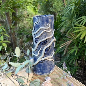 Pode incluir: Um vaso cilíndrico de cerâmica feito à mão em um tom azul escuro. O vaso apresenta linhas verticais texturizadas e é adornado com enfeites brancos, orgânicos, em forma de galho. Pequenos detalhes circulares azuis escuros também estão presentes. O vaso está sobre uma superfície de madeira.