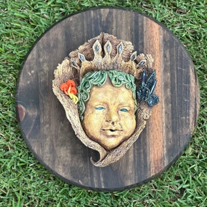 Puede incluir: Placa redonda de madera teñida oscura con una cara de niño esculpida, una corona y un marco en forma de hoja. La cara del niño es bronceada con ojos azules y cabello rizado verde. También se incluye una pequeña mariposa y setas.