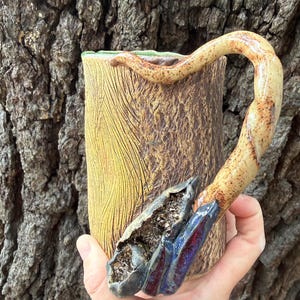 May include: A handcrafted ceramic mug with a unique design. The mug features a textured, bark-like exterior in shades of brown and yellow. The handle is a curved, branch-like shape. A decorative element with blue and red accents is on the side.
