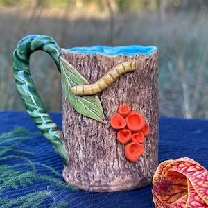 Puede incluir: Una taza de cerámica con diseño de tronco de árbol, con un asa y una hoja verdes, una oruga amarilla y detalles de hongos naranjas. El interior de la taza es azul claro.