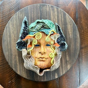 Puede incluir: Escultura de rostro de cerámica con textura de madera, adornada con hojas verdes, setas naranjas y detalles amarillos en espiral. La escultura está montada sobre una base circular de madera oscura, creando una estética natural y terrenal.