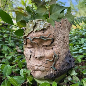 May include: A handcrafted ceramic planter shaped like a face, with closed eyes and textured details. The planter has a natural, earthy color palette with green accents and is filled with green plants, creating a unique garden decor piece.