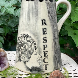 Puede incluir: Una taza de cerámica color crema con asa y borde negros. La taza presenta una ilustración en blanco y negro del rostro de una mujer y la palabra "RESPECT" en letras mayúsculas negras. La taza está sobre un fondo de follaje verde.