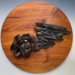 Puede incluir: Una placa de madera redonda con tinte oscuro, con una escultura de metal. La escultura representa el rostro de una mujer y un diseño abstracto. El metal tiene un acabado oscuro y envejecido, que contrasta con la madera.