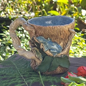 May include: A handcrafted ceramic mug with a tree trunk design, featuring a blue frog perched on a green leaf. The mug has a textured, brown exterior and a blue interior. The handle is shaped like a tree branch.