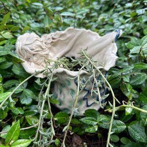 May include: A ceramic planter shaped like a reclining figure with flowing hair, nestled among green foliage. The planter is off-white with blue and green accents, and contains a trailing succulent plant. A unique decorative item for home or garden.