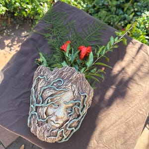 Puede incluir: Una maceta decorativa, texturizada, con aspecto de piedra, con un diseño de rostro sereno y enredaderas de color verde azulado. La maceta contiene flores rojas vibrantes y follaje verde, creando una exhibición natural y artística.