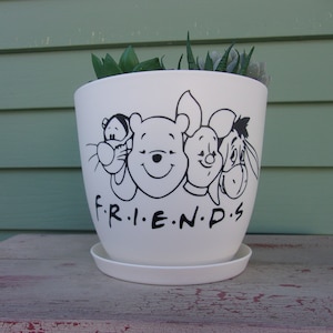 May include: White ceramic planter with a black and white graphic of Winnie the Pooh, Piglet, Tigger, and Eeyore. The planter has the words "F.R.I.E.N.D.S." printed on the front.