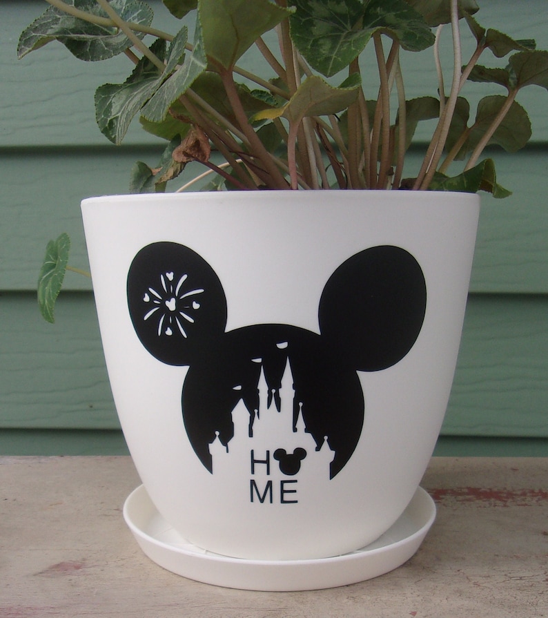 Puede incluir: Maceta de cer&aacute;mica blanca con una silueta negra de la cabeza de Mickey Mouse, el castillo de Disney y el texto "Home" con una cabeza de Mickey Mouse en lugar de la "O".