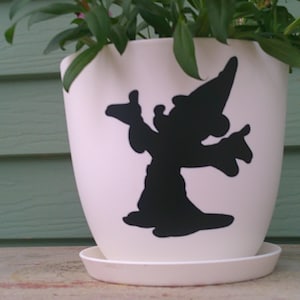 May include: A cream-colored ceramic flower pot with a black silhouette of a wizard. The pot sits on a matching saucer and contains green plants. The pot is placed in front of a green wooden wall.