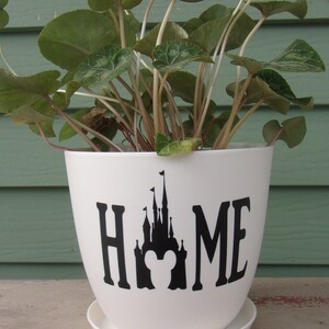 Puede incluir: Maceta de cer&aacute;mica blanca con una silueta negra del castillo de Disney y la palabra "HOME" en la parte delantera. La maceta est&aacute; llena de plantas verdes de hoja.