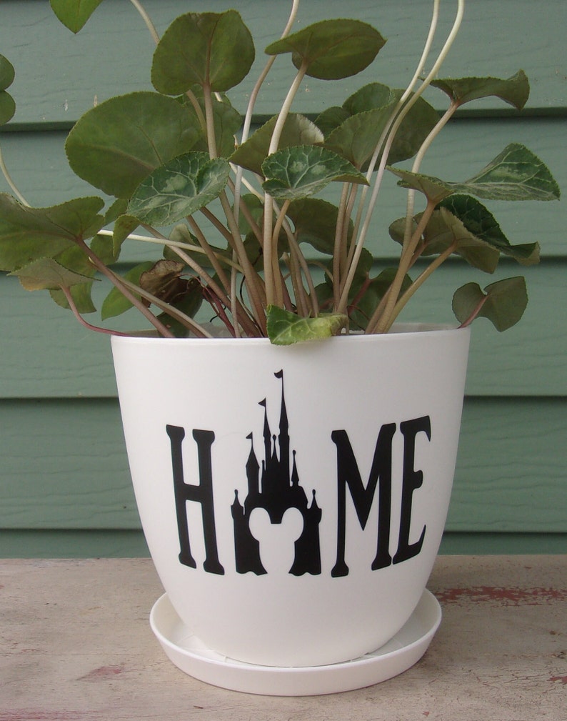 Puede incluir: Maceta de cer&aacute;mica blanca con una silueta negra del castillo de Disney y la palabra "HOME" en la parte delantera. La maceta est&aacute; llena de plantas verdes de hoja.