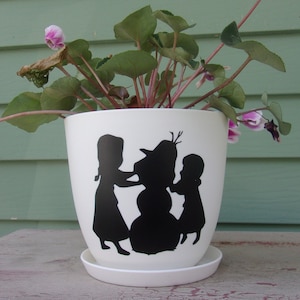 May include: White ceramic planter with a black silhouette of two children and a snowman. The planter has a green plant with pink flowers growing in it.