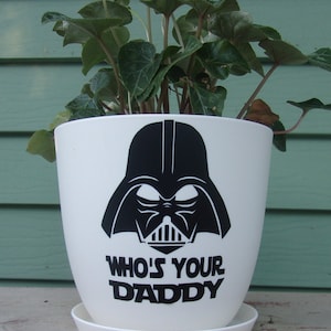 May include: White ceramic planter with a black Darth Vader graphic and the text "Who's Your Daddy". The planter is filled with green leafy plants.