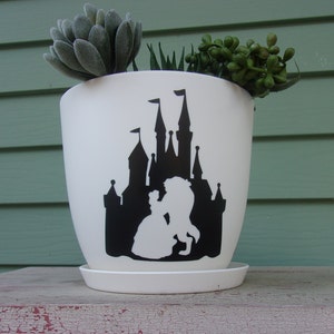 May include: White ceramic planter with a black silhouette of Beauty and the Beast in front of a castle. The planter has a matching saucer.