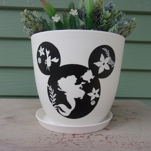 May include: White ceramic planter with a black silhouette of Ariel from The Little Mermaid, Sebastian, Flounder, and other sea creatures. The planter is shaped like Mickey Mouse's head.