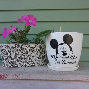 May include: White ceramic flower pot with a black and white Mickey Mouse design. The pot has a saucer and the text "The Ormans" is printed on the front.