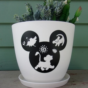 May include: White ceramic planter with a Mickey Mouse silhouette design featuring characters from The Lion King. The planter has a black and white silhouette of Simba, Pumbaa, Timon, and the sun. The planter has a saucer.