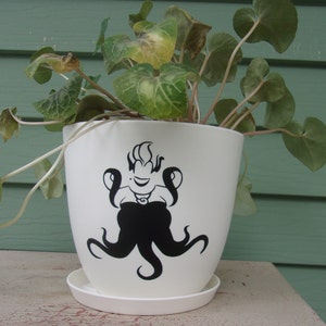 May include: White ceramic planter with a black and white illustration of a cartoon octopus wearing a crown. The planter is filled with green leafy plants.