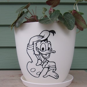 May include: White ceramic planter with a black and white illustration of a cartoon duck wearing a hat. The planter has a saucer and is filled with green leafy plants.