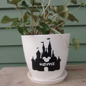 Puede incluir: Maceta de cer&aacute;mica blanca con una silueta negra del castillo de Disney y las orejas de Mickey Mouse con la palabra "HOME" debajo. La maceta est&aacute; llena de una planta verde.