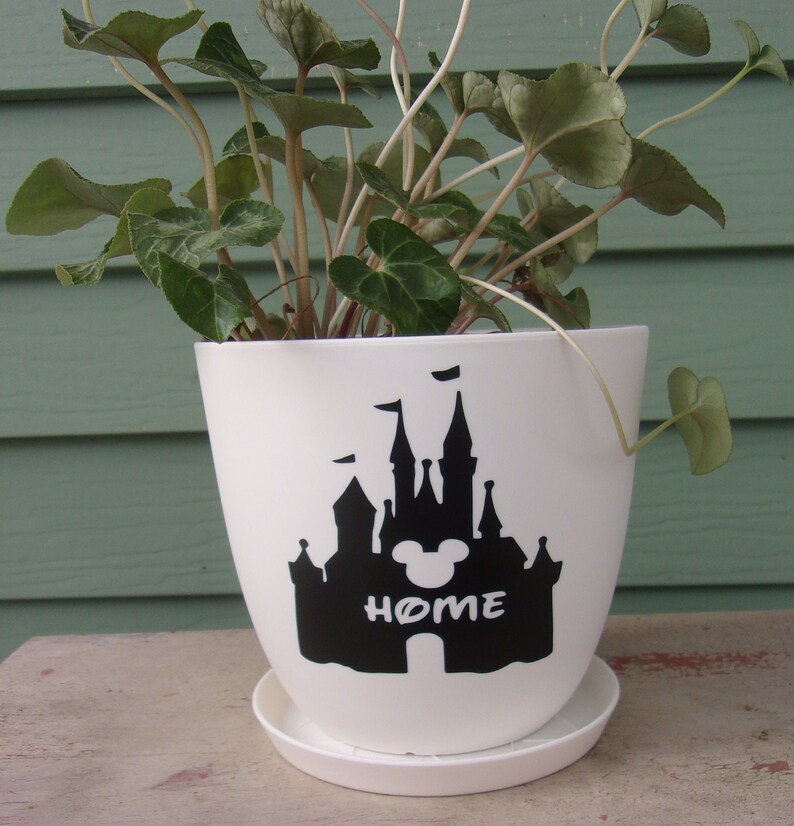 Puede incluir: Maceta de cer&aacute;mica blanca con una silueta negra del castillo de Disney y las orejas de Mickey Mouse con la palabra "HOME" debajo. La maceta est&aacute; llena de una planta verde.
