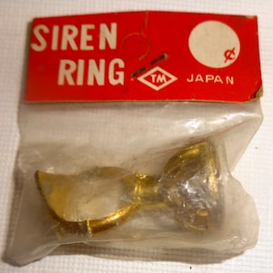 May include: A vintage Siren Ring in a clear plastic bag with a red label. The label reads "SIREN RING" and "JAPAN" in white text. The ring is gold-colored.