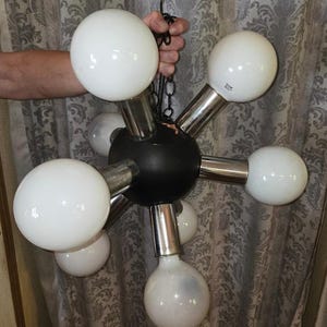 May include: A mid-century modern chandelier with a black central sphere and chrome arms, each holding a large, round, white light bulb. The fixture is suspended by a chain and is held by a hand.
