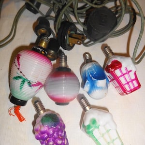 May include: A collection of vintage Christmas light bulbs in various shapes, including a grape cluster, ice cream cone, and lantern. The bulbs are multicolored and connected by a green cord with a black plug.