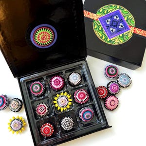May include: A black gift box with an open lid revealing a tray of colorful, circular decorative items. The items are in various colors, including pink, yellow, and blue. A second box is visible with a decorative design on the lid.