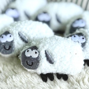 May include: A collection of white plush sheep toys with embroidered black and gray faces and black hooves. The sheep have a fluffy, textured appearance and are arranged on a soft, white surface.