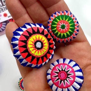 May include: Three circular embroidered buttons featuring intricate geometric patterns in vibrant neon pink, yellow, green, white, and royal blue thread. Each button displays a sunburst or mandala-like design with radiating spikes and concentric circles.