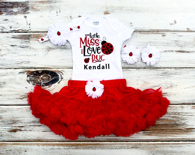 May include: White bodysuit with red ladybug print and the text "Little Miss Love Bug Kendall". Red tulle skirt with white flower embellishment. Red ladybug headband.