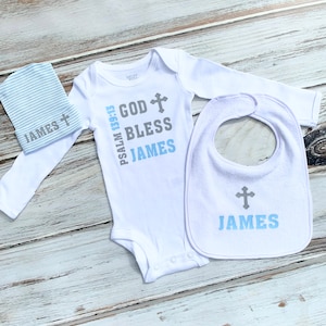 K&ouml;nnte beinhalten: Ein wei&szlig;er Baby-Strampler mit langen &Auml;rmeln, ein L&auml;tzchen und eine M&uuml;tze. Der Strampler hat den Text "PSALM 139:13 GOD BLESS JAMES" mit einem Kreuzsymbol. Das L&auml;tzchen und die M&uuml;tze haben den Text "JAMES" mit einem Kreuzsymbol.
