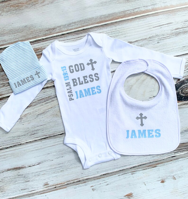 K&ouml;nnte beinhalten: Ein wei&szlig;er Baby-Strampler mit langen &Auml;rmeln, ein L&auml;tzchen und eine M&uuml;tze. Der Strampler hat den Text "PSALM 139:13 GOD BLESS JAMES" mit einem Kreuzsymbol. Das L&auml;tzchen und die M&uuml;tze haben den Text "JAMES" mit einem Kreuzsymbol.
