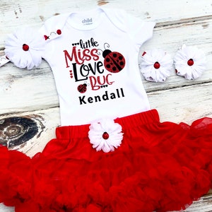 May include: A white bodysuit with a red ladybug design and the text "little Miss Love Bug Kendall". The outfit includes a red tulle skirt with a white flower and a white headband with a red ladybug.