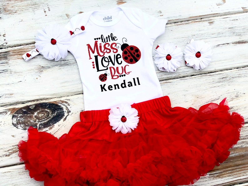 May include: A white bodysuit with a red ladybug design and the text "little Miss Love Bug Kendall". The outfit includes a red tulle skirt with a white flower and a white headband with a red ladybug.