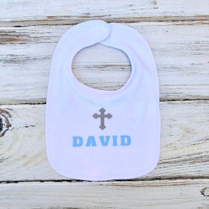 K&ouml;nnte beinhalten: Wei&szlig;er Baby-L&auml;tzchen mit einem grauen Kreuz und dem Namen "DAVID" in blauen Buchstaben.