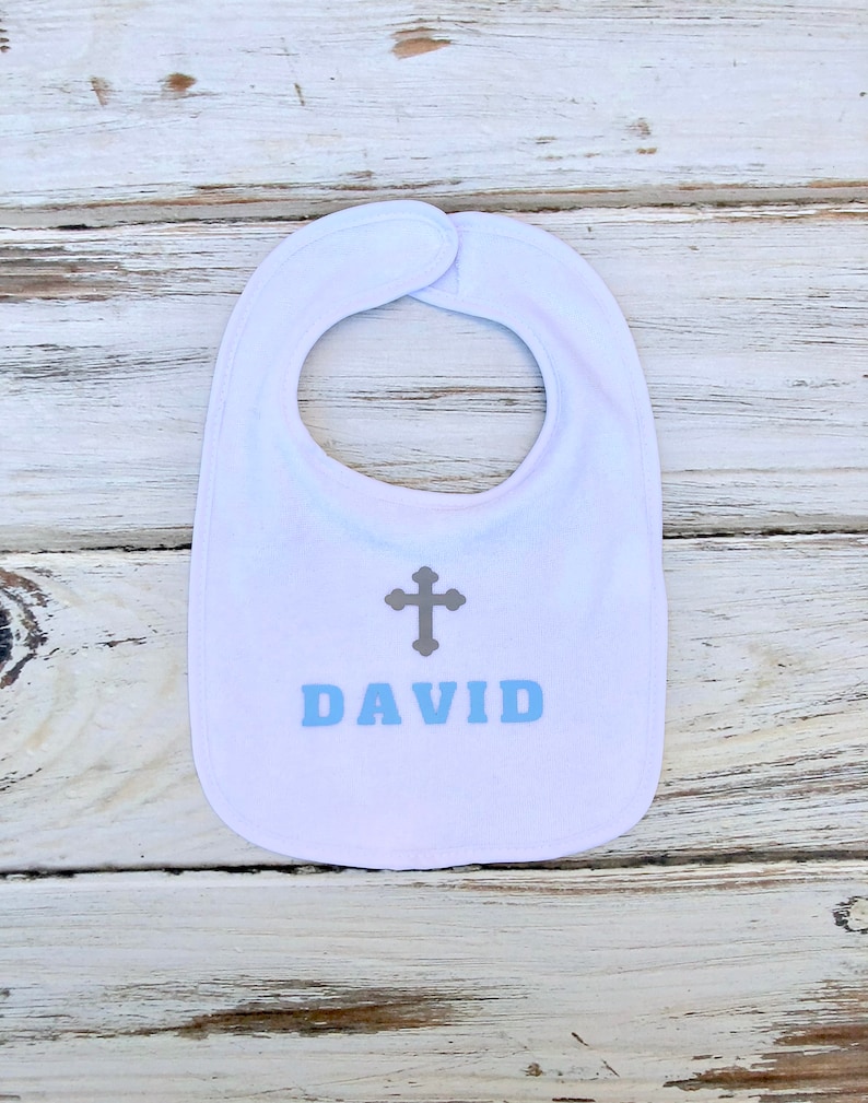 K&ouml;nnte beinhalten: Wei&szlig;er Baby-L&auml;tzchen mit einem grauen Kreuz und dem Namen "DAVID" in blauen Buchstaben.