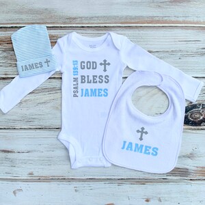 K&ouml;nnte beinhalten: Ein wei&szlig;es Baby-Body, L&auml;tzchen und M&uuml;tzen-Set mit einem grauen Kreuz und dem Text "God Bless James" und "Psalm 139:13".