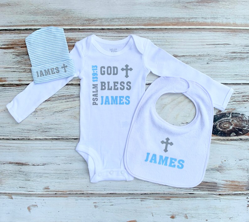K&ouml;nnte beinhalten: Ein wei&szlig;es Baby-Body, L&auml;tzchen und M&uuml;tzen-Set mit einem grauen Kreuz und dem Text "God Bless James" und "Psalm 139:13".