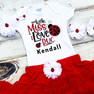 May include: A white bodysuit with a red and black ladybug design and the text "little Miss Love Bug Kendall". The outfit includes a red tulle skirt with a white flower and a white headband with a red ladybug.